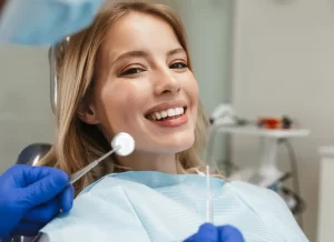 A young woman sitting in a dental chair, smiling while a dentist holds up a dental mirror for her exam.